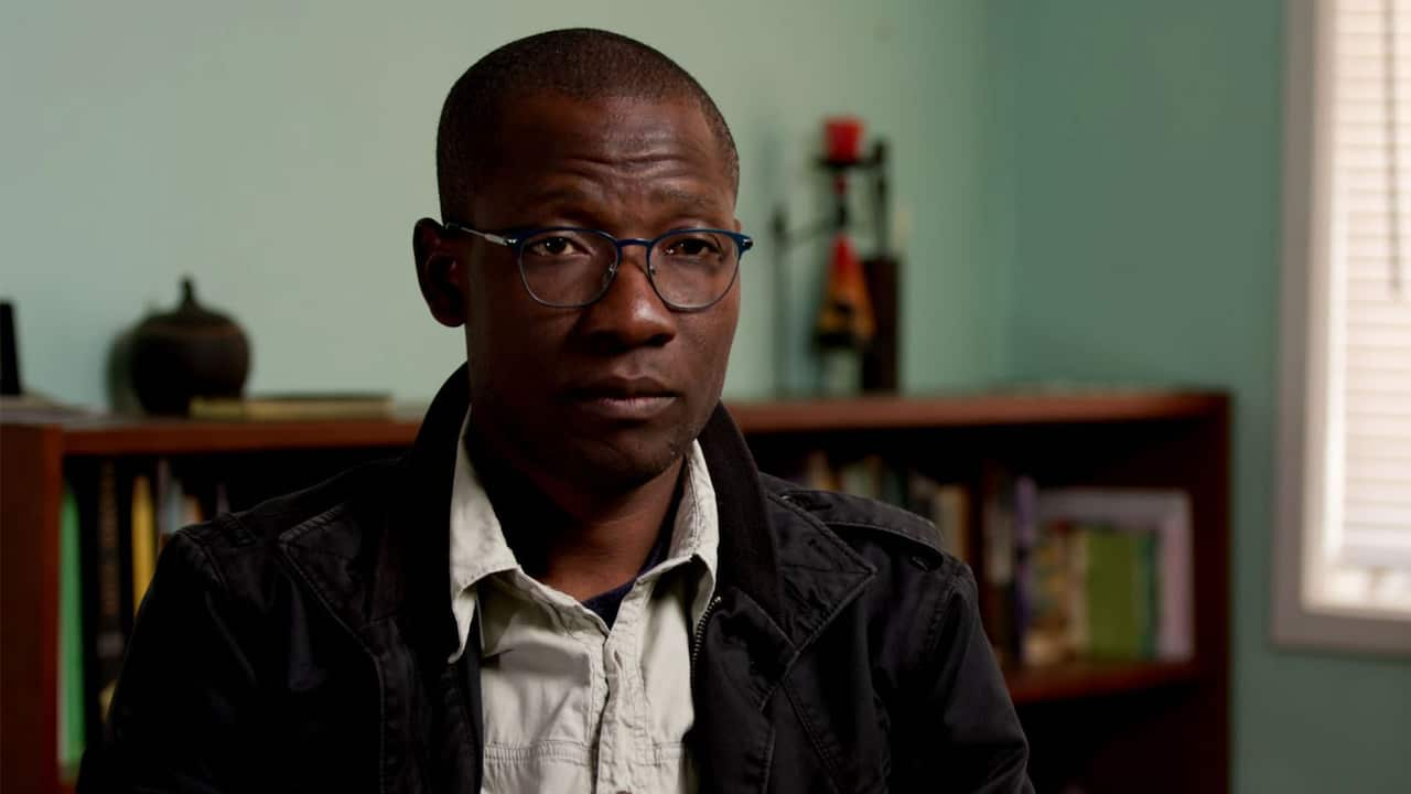 An African man wearing prescription glasses, a white shirt and a black jacket 