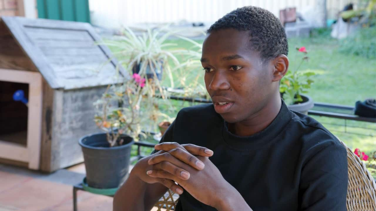 An African teenage boy wearing a black sweater is speaking with his fingers interlaced 