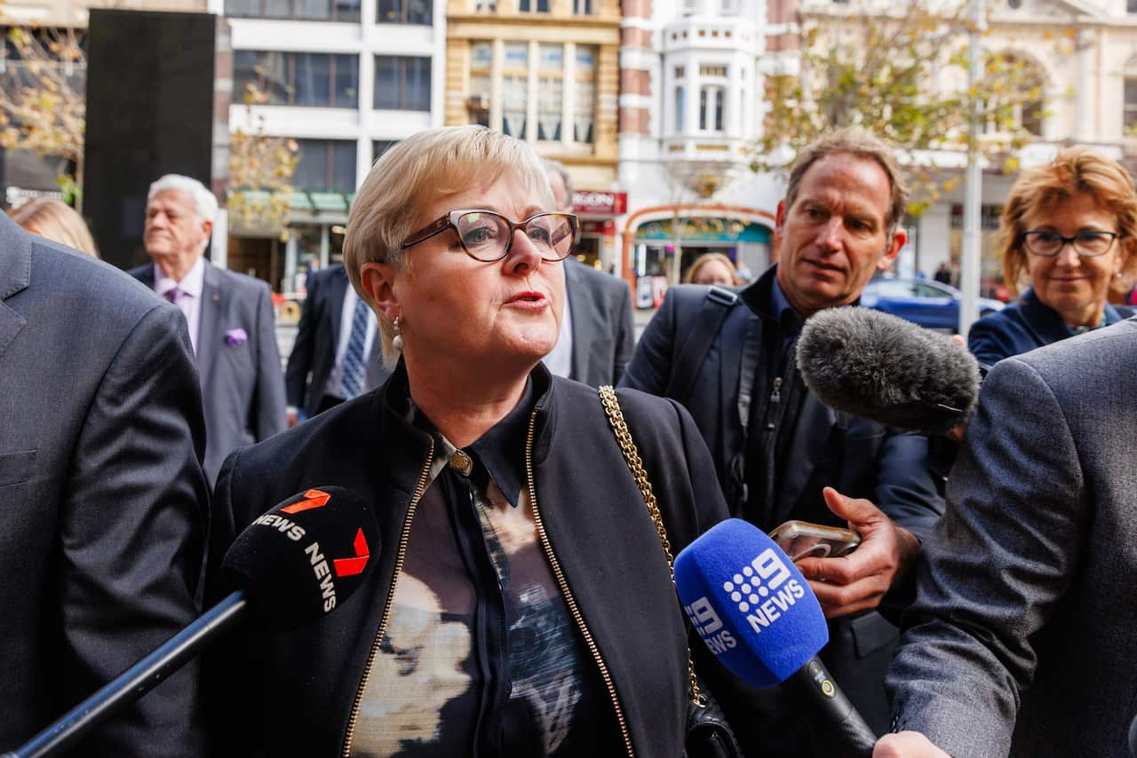 A woman in a black jacket (Linda Reynolds) is swarmed by journalists carrying microphones outside a courthouse. 