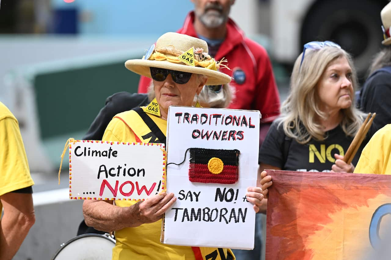 A woman holding signs that read 'Climate Action NOW' and 'Traditional owners say no to Tamboran' with an Indigenous flag on it.