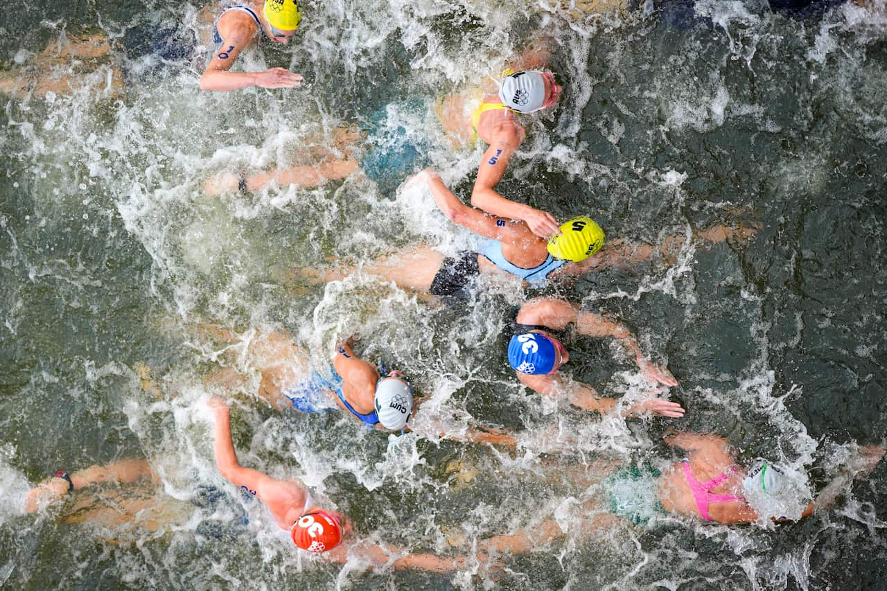 Several swimmers wearing differently coloured caps, swimming in the water and creating waves. 