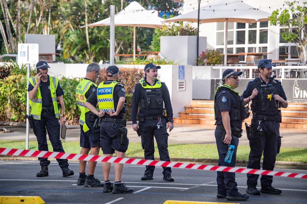 Several police standing behind white and red tape. A white hotel is in the background.