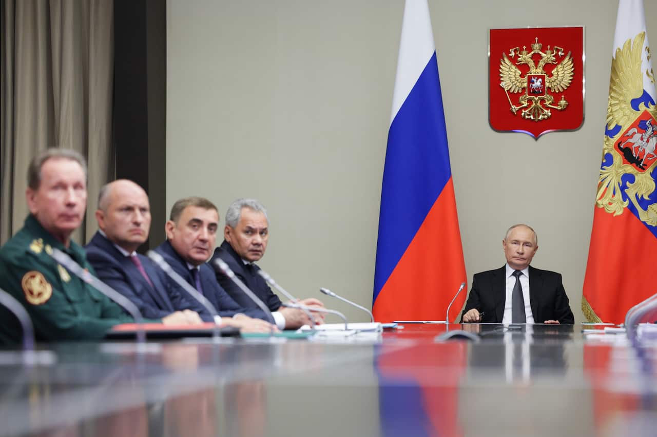 A man in a suit sits behind a desk, military men are in a meeting with him