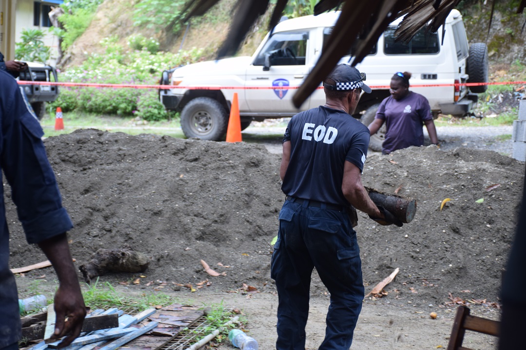 An officer removes an unexploded bomb from a site. There is a large mound of dirt in front of him. 