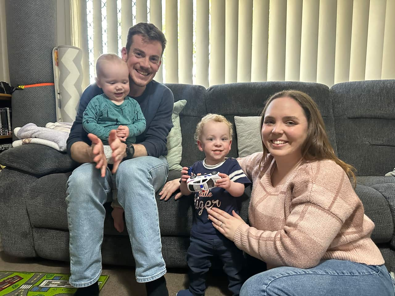 A family posing for a photo. The father is sitting on a couch with his child seated on his lap. The mother is seated on the floor leaning against the couch with her arm over the shoulders of another of their children who is standing next to her.