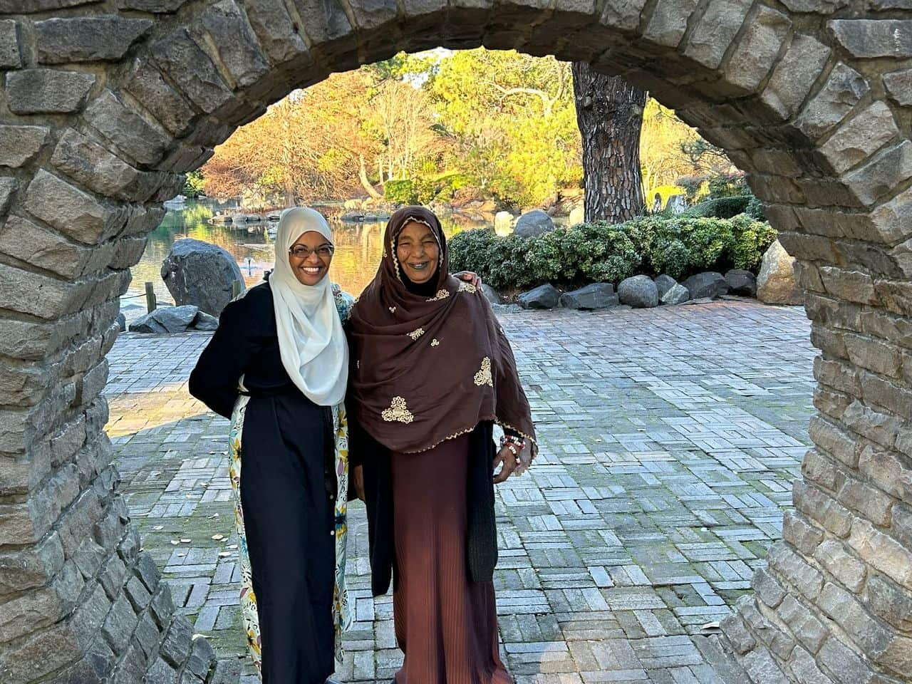 Two women wearing headscarves and standing outside smiling and looking towards the camera