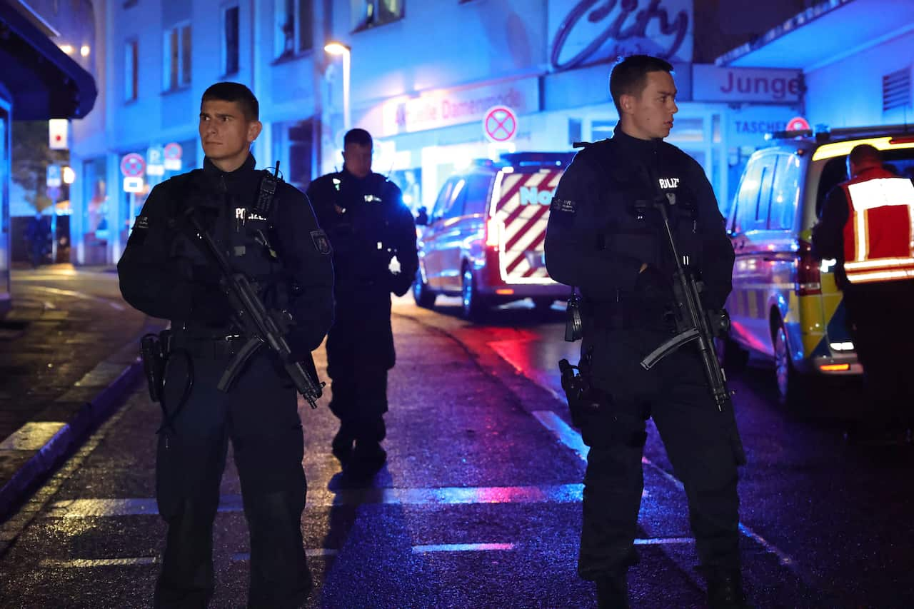 Two police with guns standing on a road, with ambulances behind them to the side.