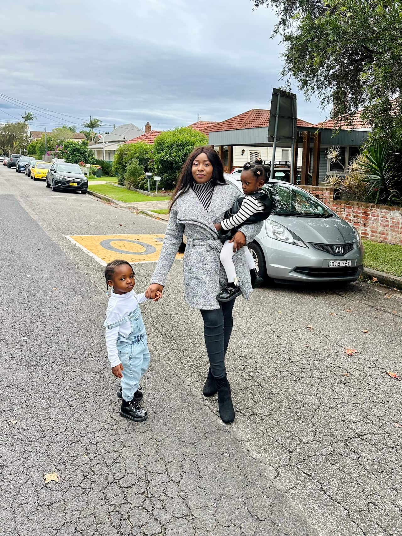 A woman walks along a suburban street while holding one child in her arms and another's hand.