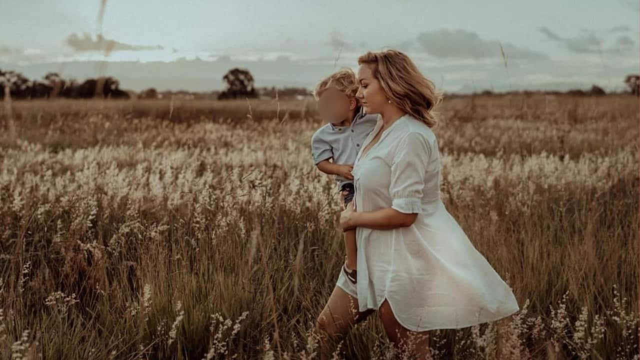 A woman walking through a field of crops carrying a child whose face is blurred.