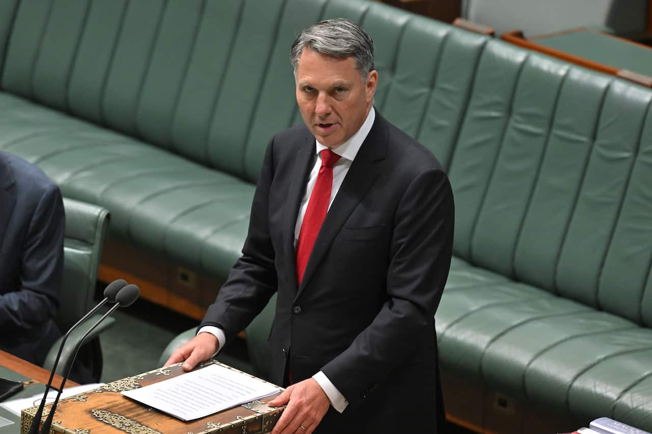 A man stands at a lectern in a suit, reading a speech in Parliament.