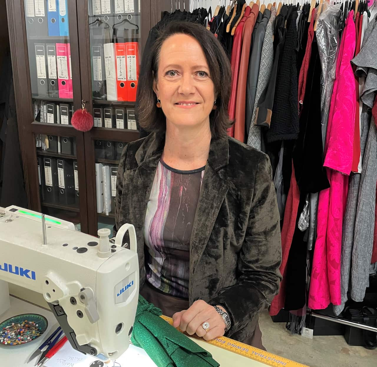 A woman in a brown jacket sits at a sewing machine in a fashion studio.