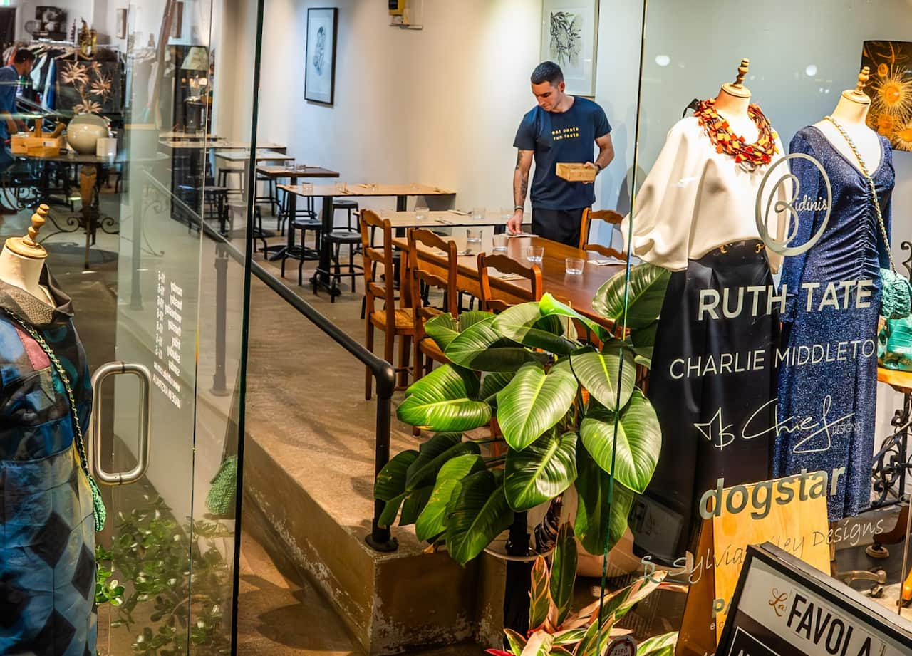 The view through a glass window shows restaurant tables being set up inside a fashion store. 