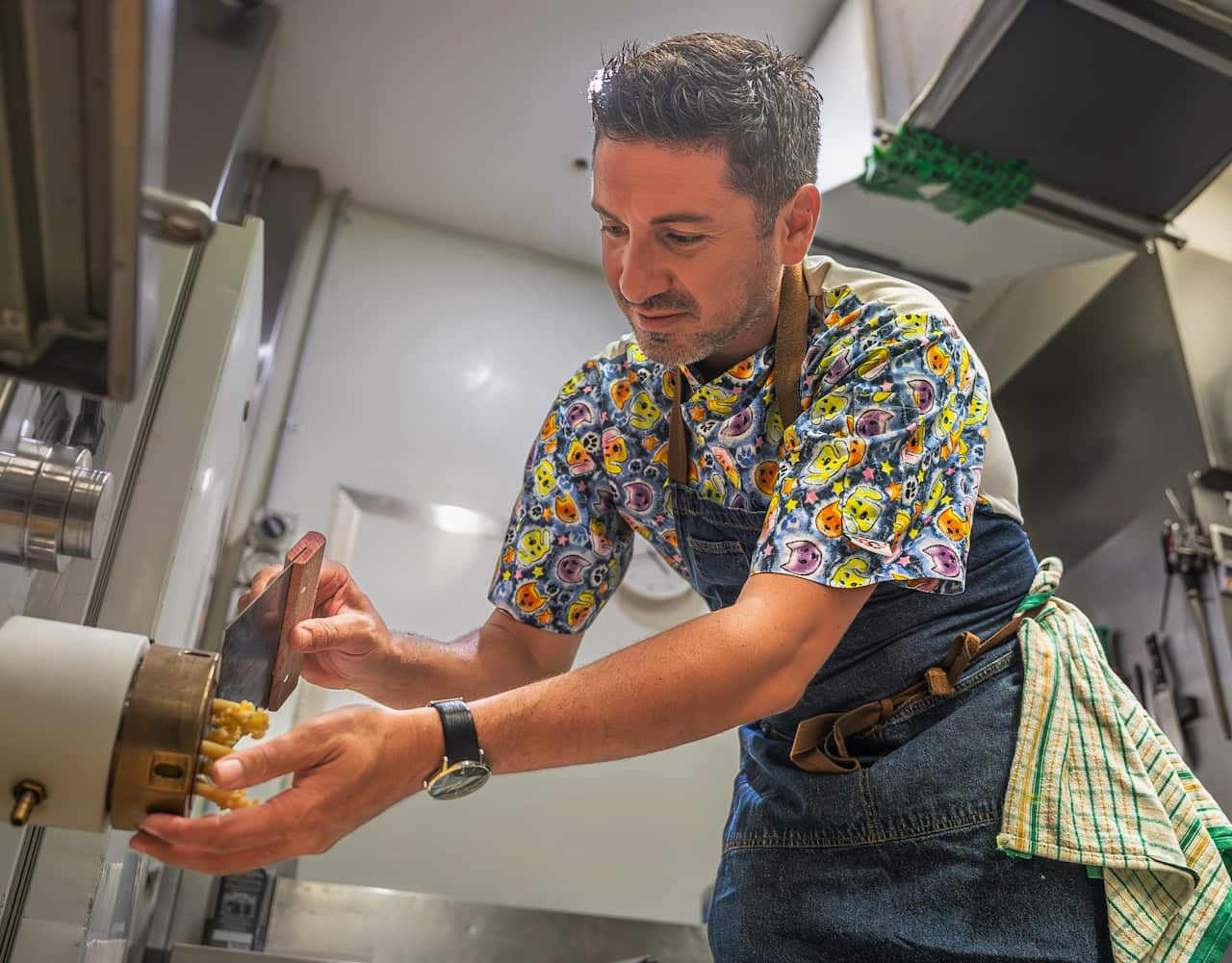 A man in a colourful shirt extrudes pasta from a small device.