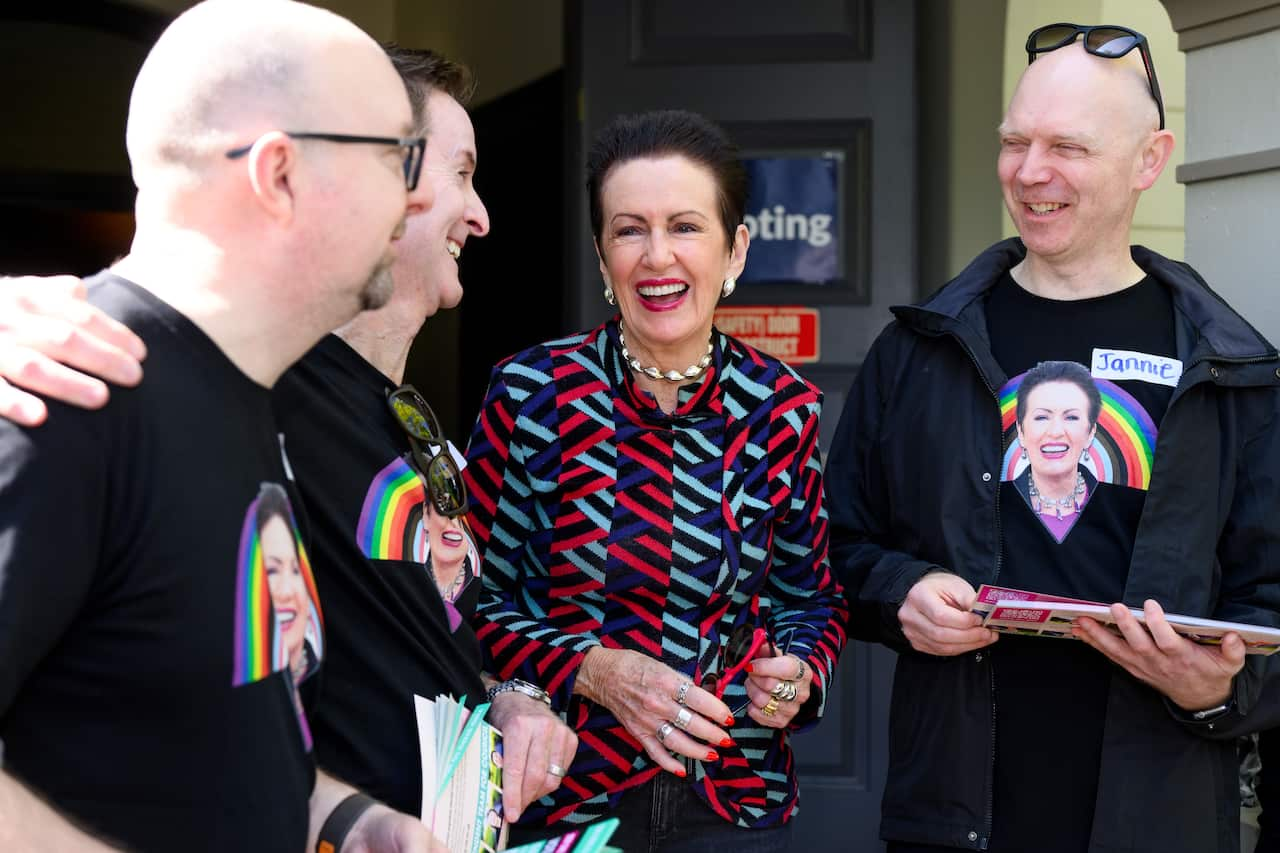 A woman with tied-back dark hair and a teal, red and dark blue patterned shirt stands between several men wearing T-shirts emblazoned with a picture of her face and a rainbow. 