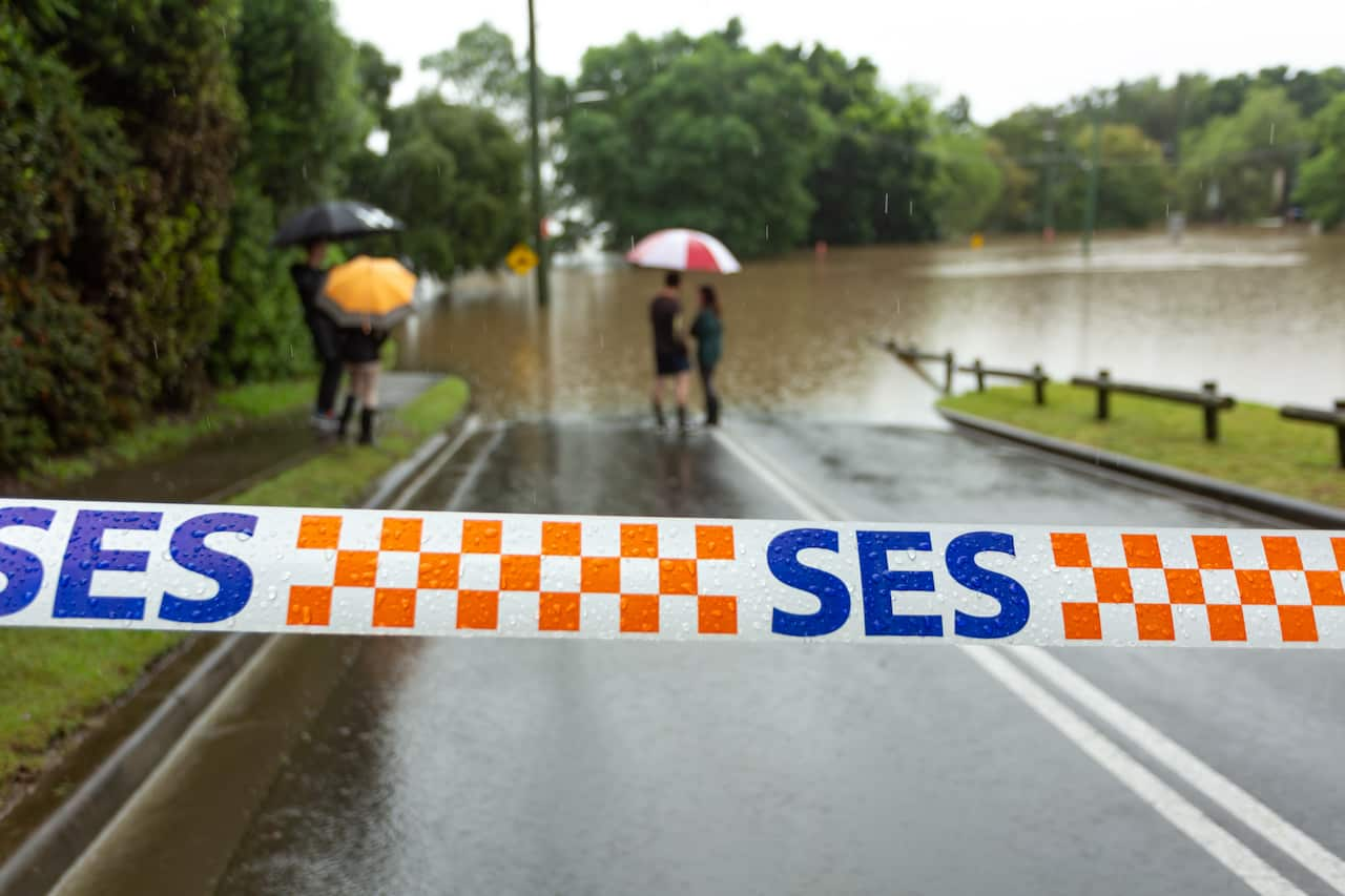 SES tape blocks off a road, where people are standing near a flood site and holding umbrellas.