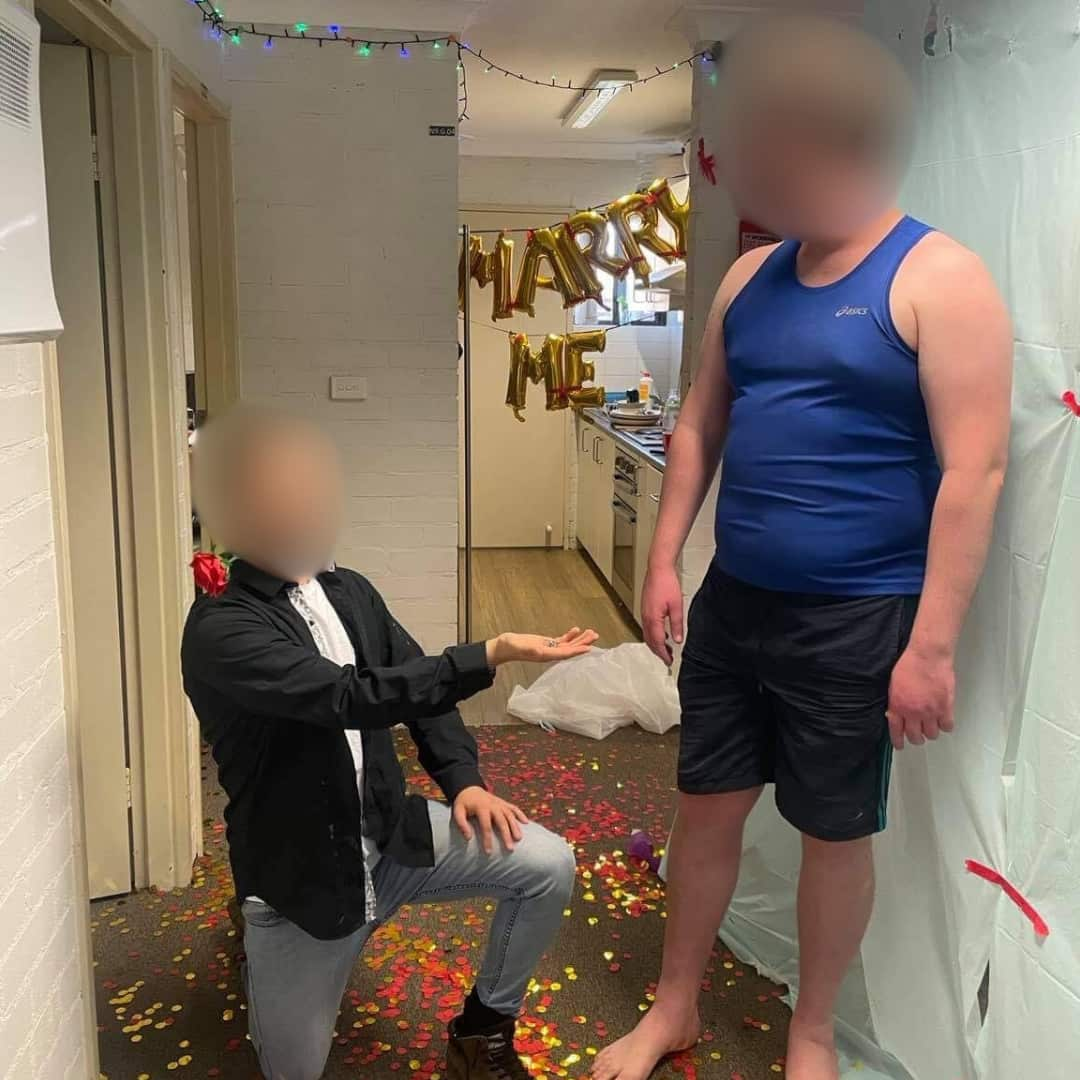 A man with his face blurred kneeling on the ground, reaching out his hand to a standing man with his face blurred. Confetti is scattered around them and gold balloons spelling out Marry Me are behind them.