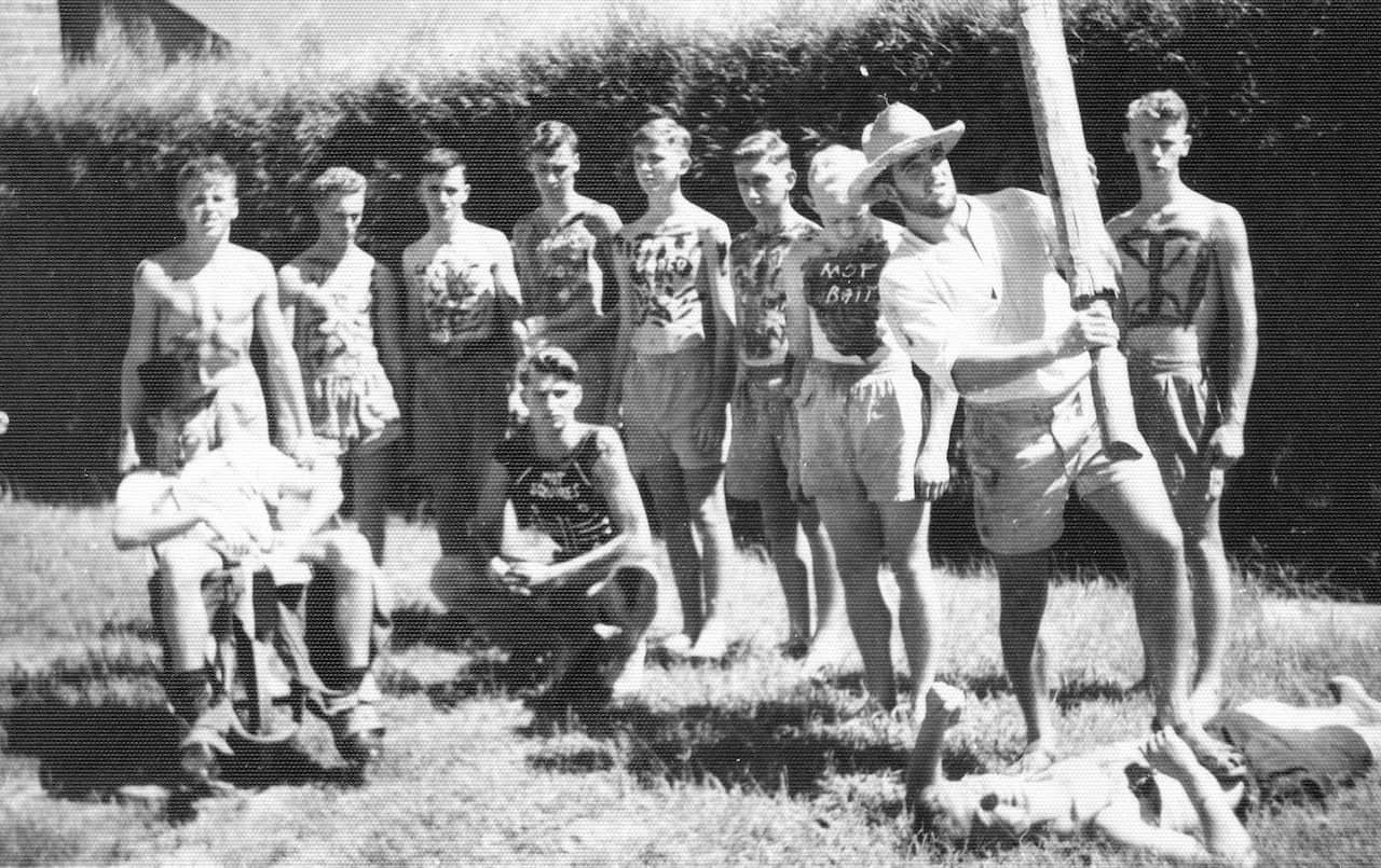 A black-and-white photo of 11 young men in shorts. Ten are shirtless and many have words and designs painted on their chest like "mot bait", while the other hold a large wooden pole