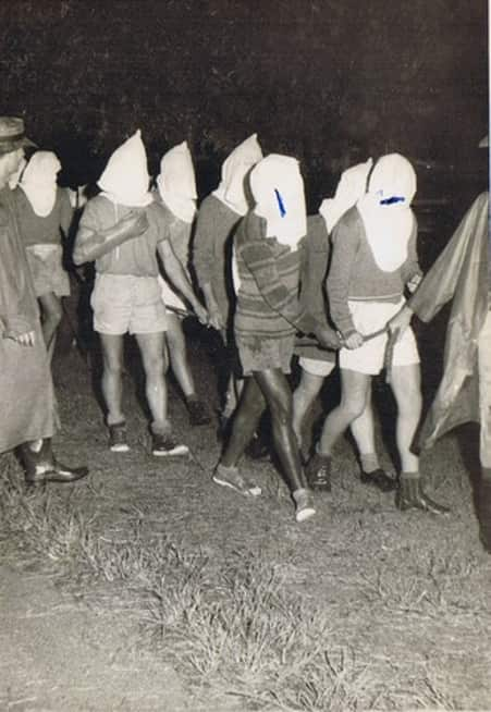 A black-and-white image of a group of young men walking with white sacks covering their faces