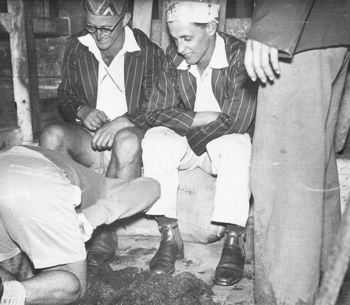 A black-and-white image of a man kneeling with his head to the ground in front of two men in pinstriped blazers wearing crowns