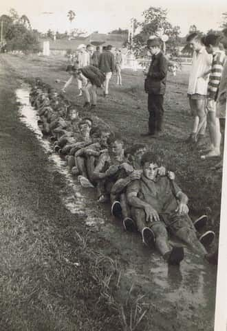 A black-and-white image of a group of young men sitting down in a line, grasping each other's shoulders and covered in mud while other students look on