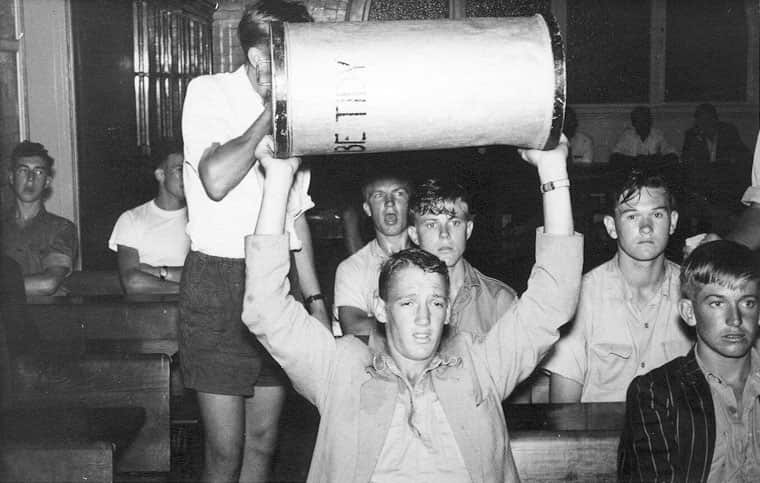 A black-and-white image of students in a classroom. A young man in the front holds a large, metal rubbish bin above his head