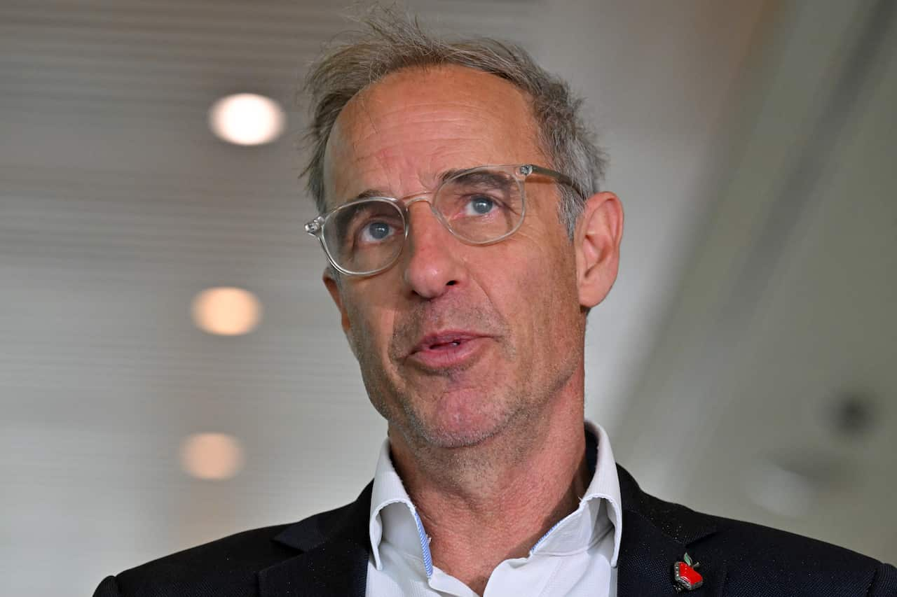 A man with short grey hair and clear glasses wearing a navy blazer, white shirt, and apple-shaped pin.