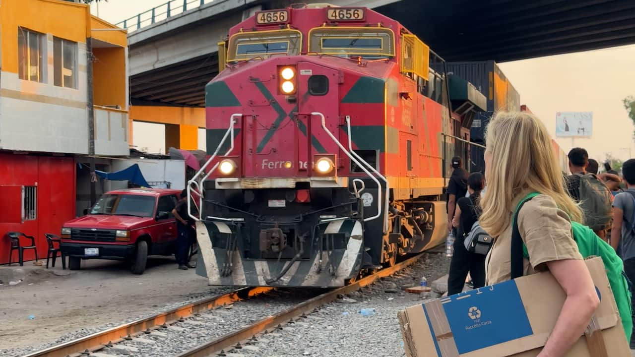 A woman with blond hair holding a cardboard box looks towards a red locomotive.