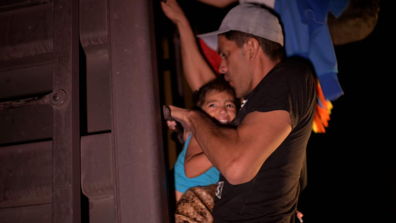 A frightened child is gripped in the hands of a man in a black T-shirt holding onto the train.