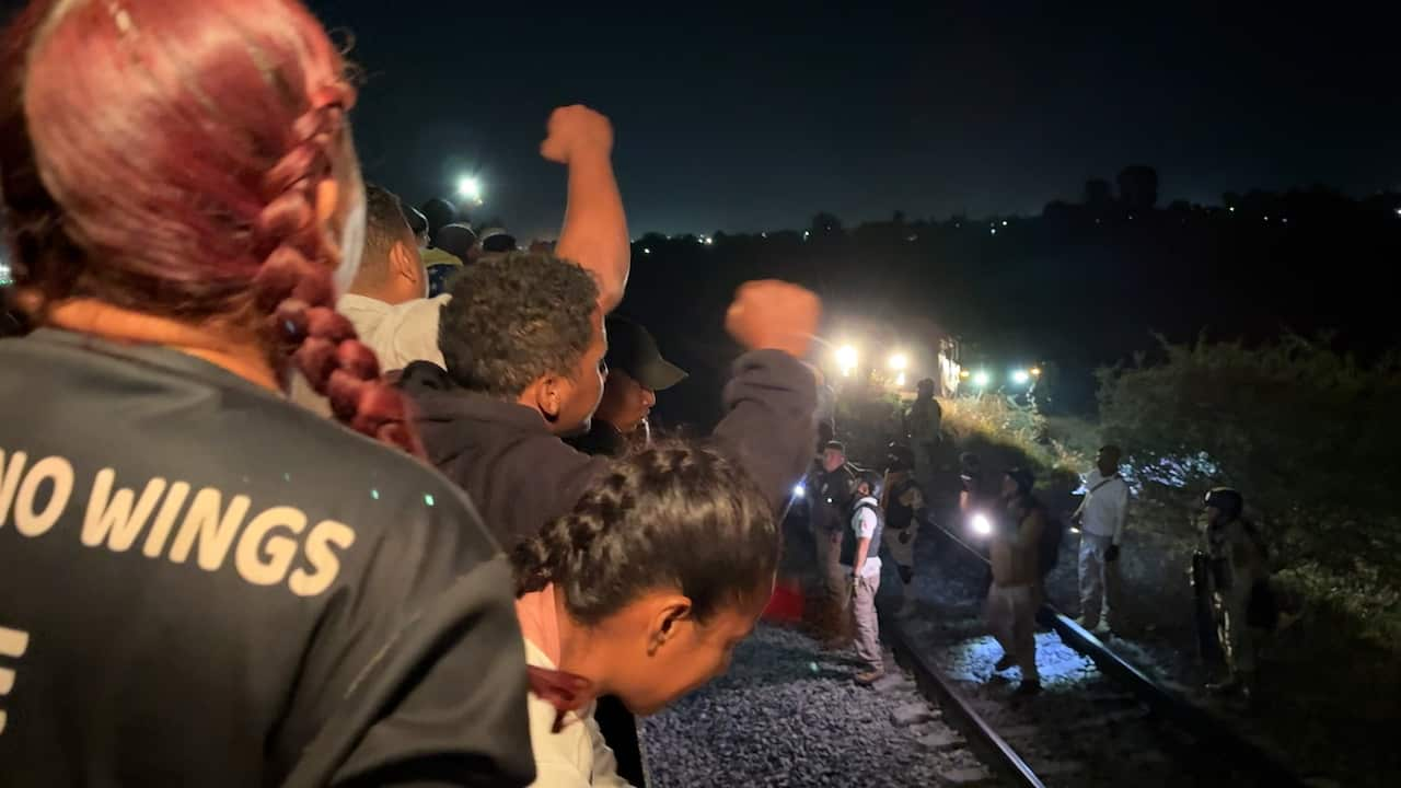 Migration authorities on the tracks in black vests shine torches up at the migrants waving their fists.