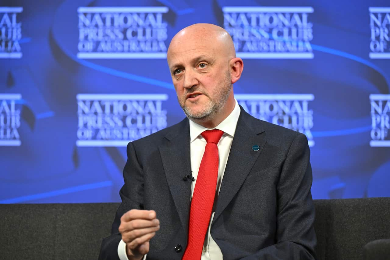 A man in a dark suit and red tie is speaking.