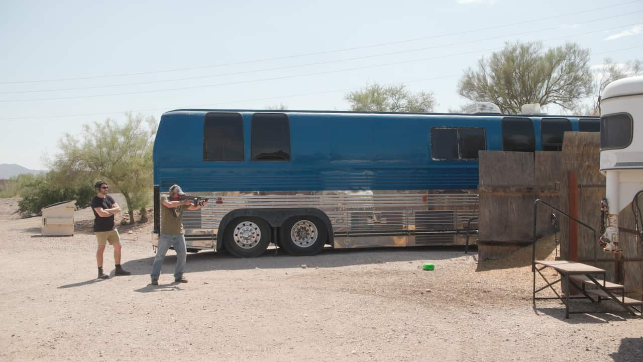 Jerry Sheridan in jeans and green shirt fires a gun at a range. He stands before a large blue bus. Dateline's Darren Mara watches on wearing ear protection.