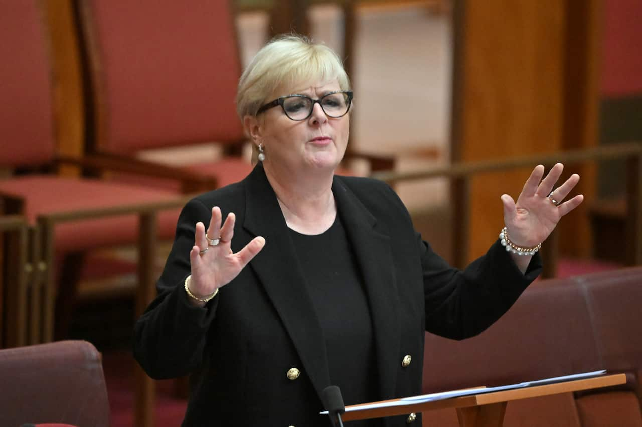 A woman in a black blazer is speaking at a lectern with her hands extended.