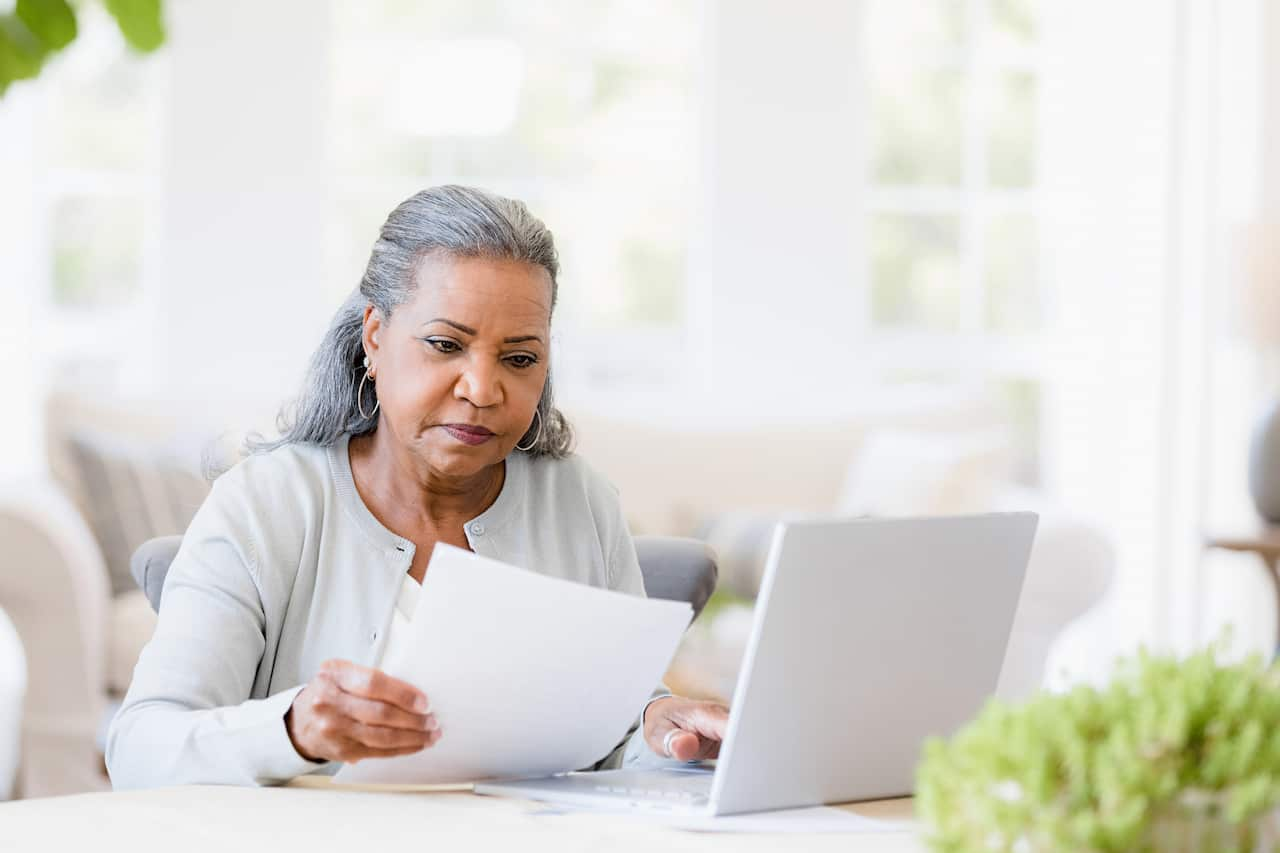 A middle-aged woman sitting at desk with a computer looking at some paperwork with a serious expression on her face.