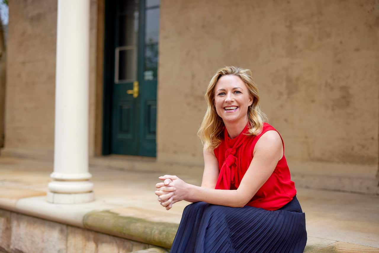 A woman wearing a blue skirt and red top sitting on a step with her hands together in front of her.