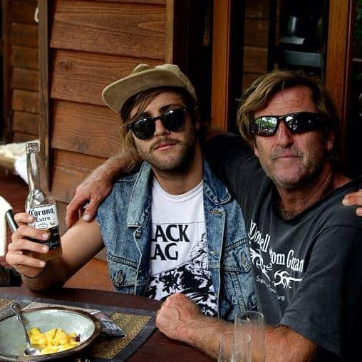 Two men wearing sunglasses sitting at a table with arms around each other.