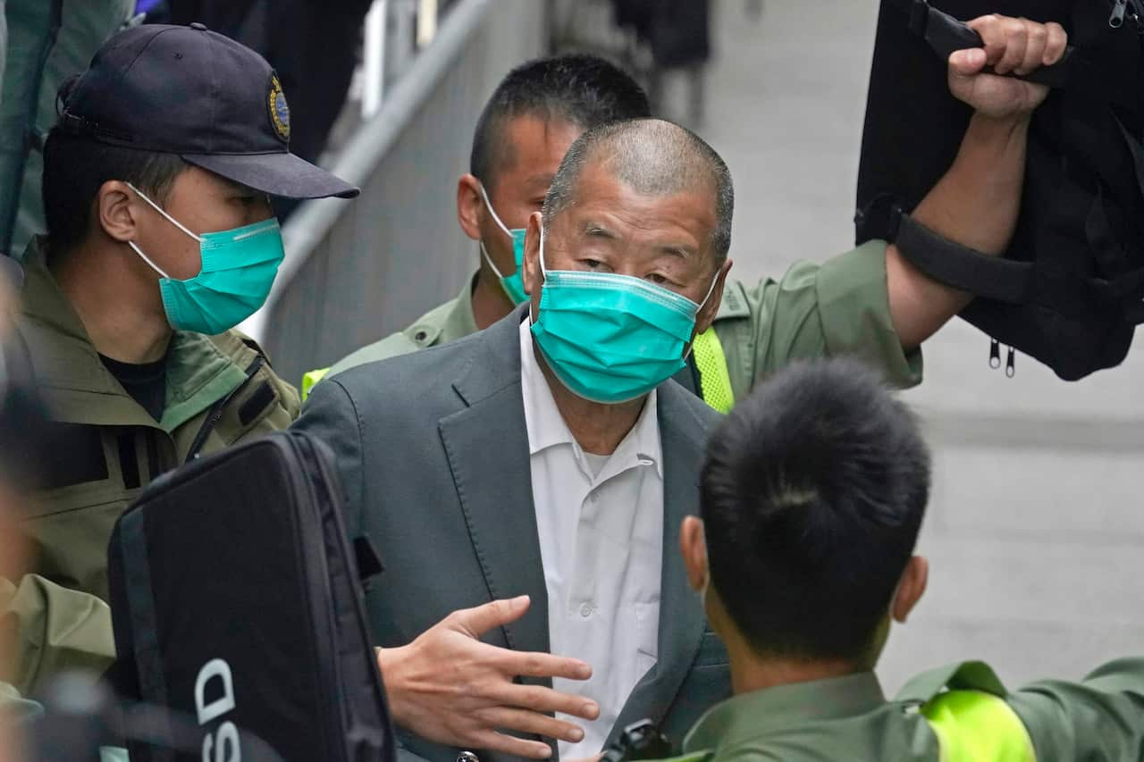 A man in a suit wearing a mask is surrounded by police officers.