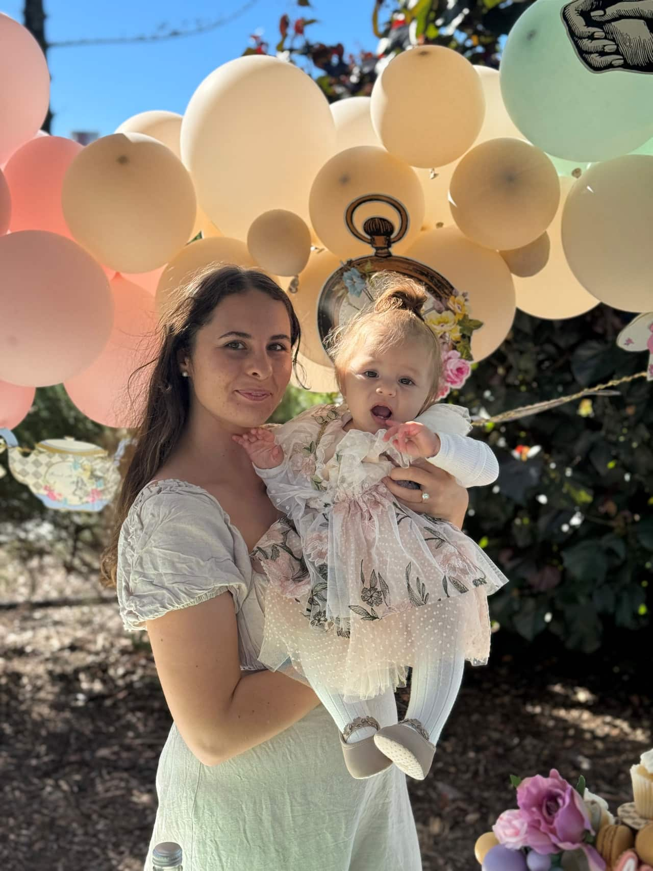 A woman in a light green dress holding a baby with balloons in the background.