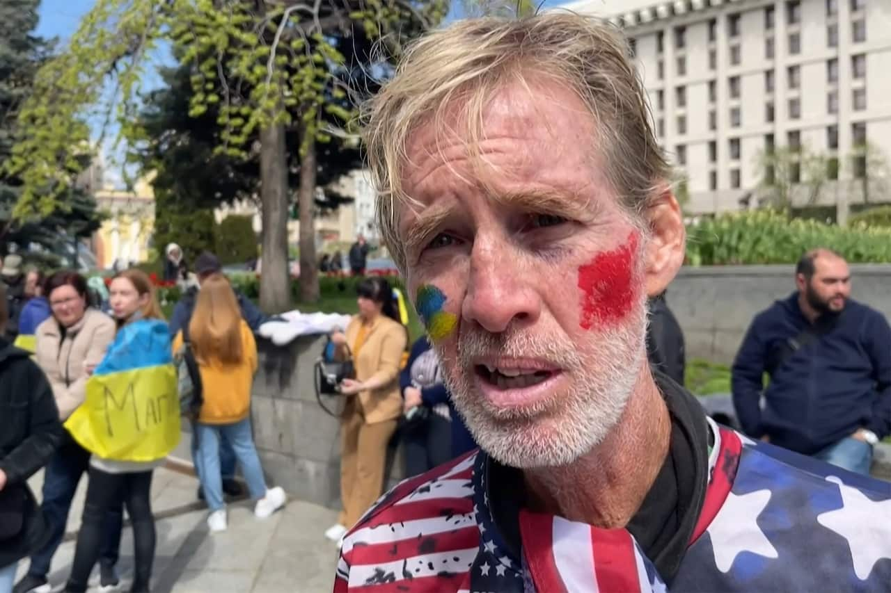 A man wearing a top featuring the US flag standing outside. He has yellow and blue paint on his right cheek and a square of red paint on his left cheek.