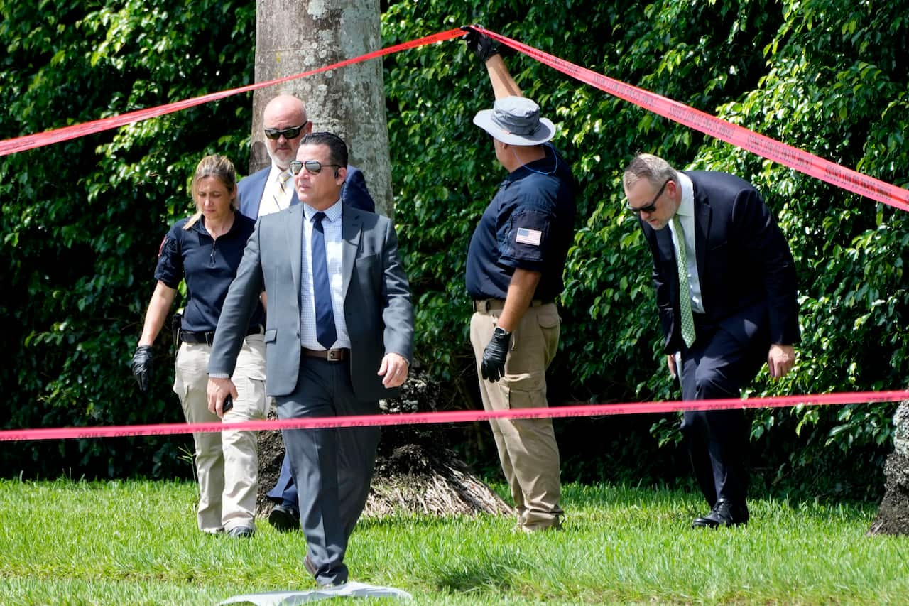 Four men and a woman on an area of grass near bushes. One man is holding up red police tape so another man can step under it