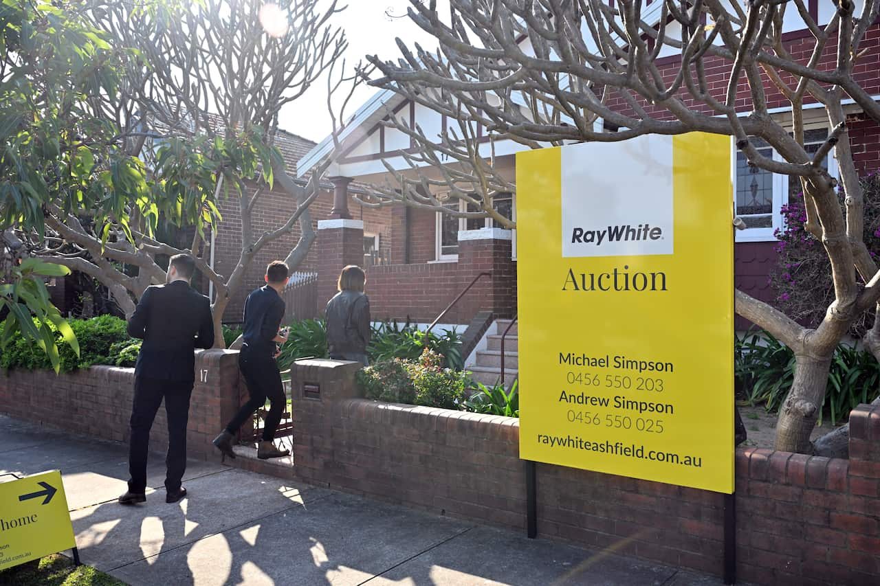 Three people standing outside a house with a sign that says Auction