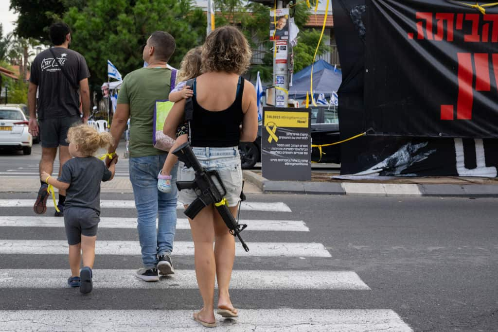 A woman walking on a road, carrying a child, with an assault rifle on her back