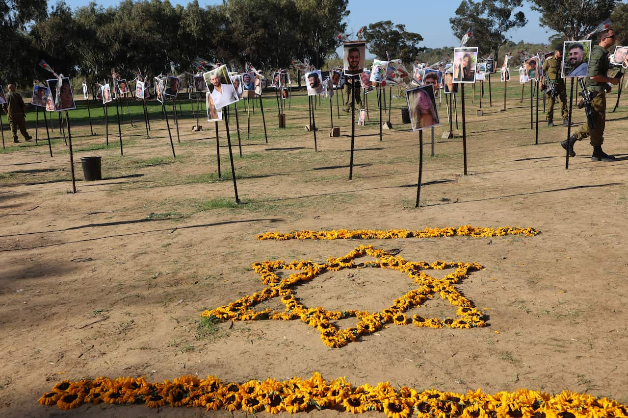 Sunflowers are arranged in the shape of an Israeli flag. In the background are many poles with photos of people attached.