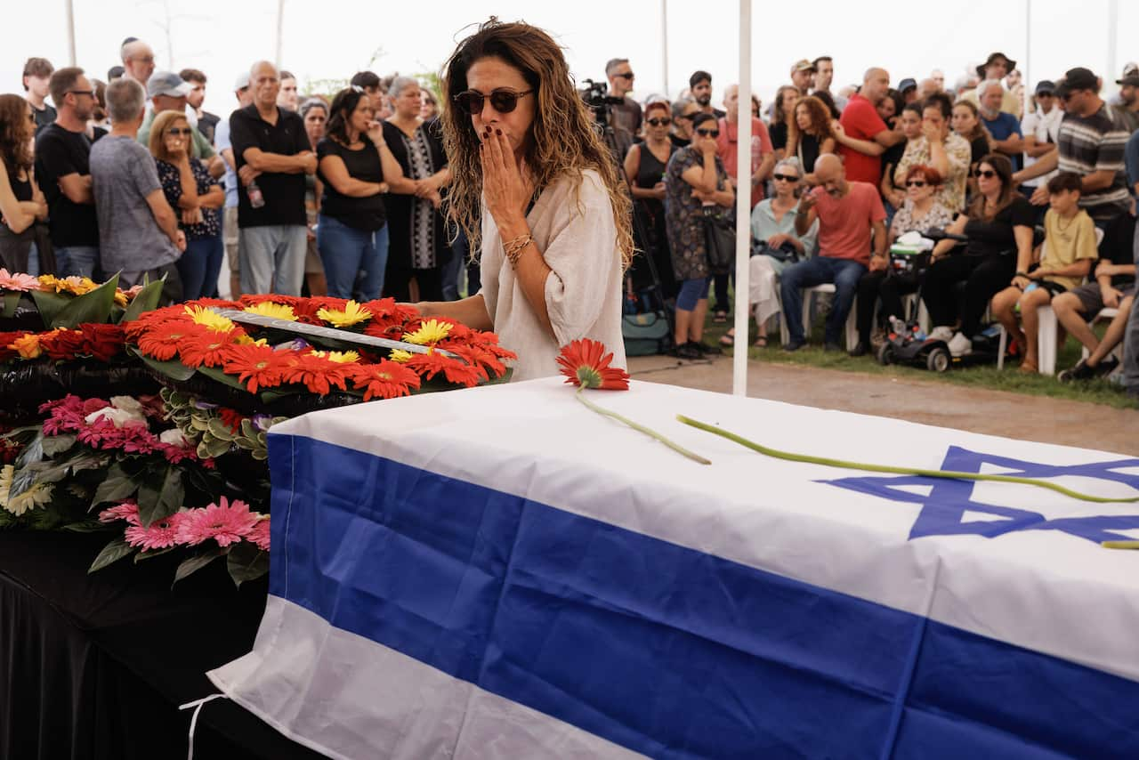 A woman wearing sunglasses holds her hand to her mouth as she stands over a coffin draped in the Israeli flag.