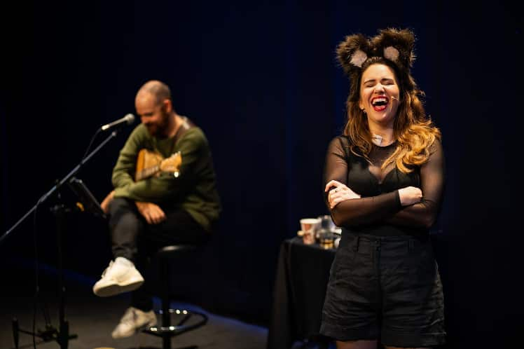 A woman in the foreground laughing while wearing a bear ear handband and black top and shorts. A man is sitting on a stool in the background holding a guitar. 