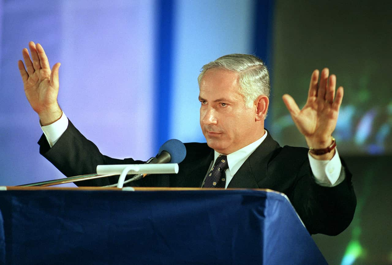 A man in a suit stands before a lectern, raising his hands.