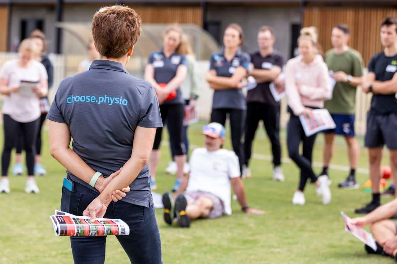 A woman stands facing a group of people on an area of grass with her back to the camera. She wears a sports polo shirt.